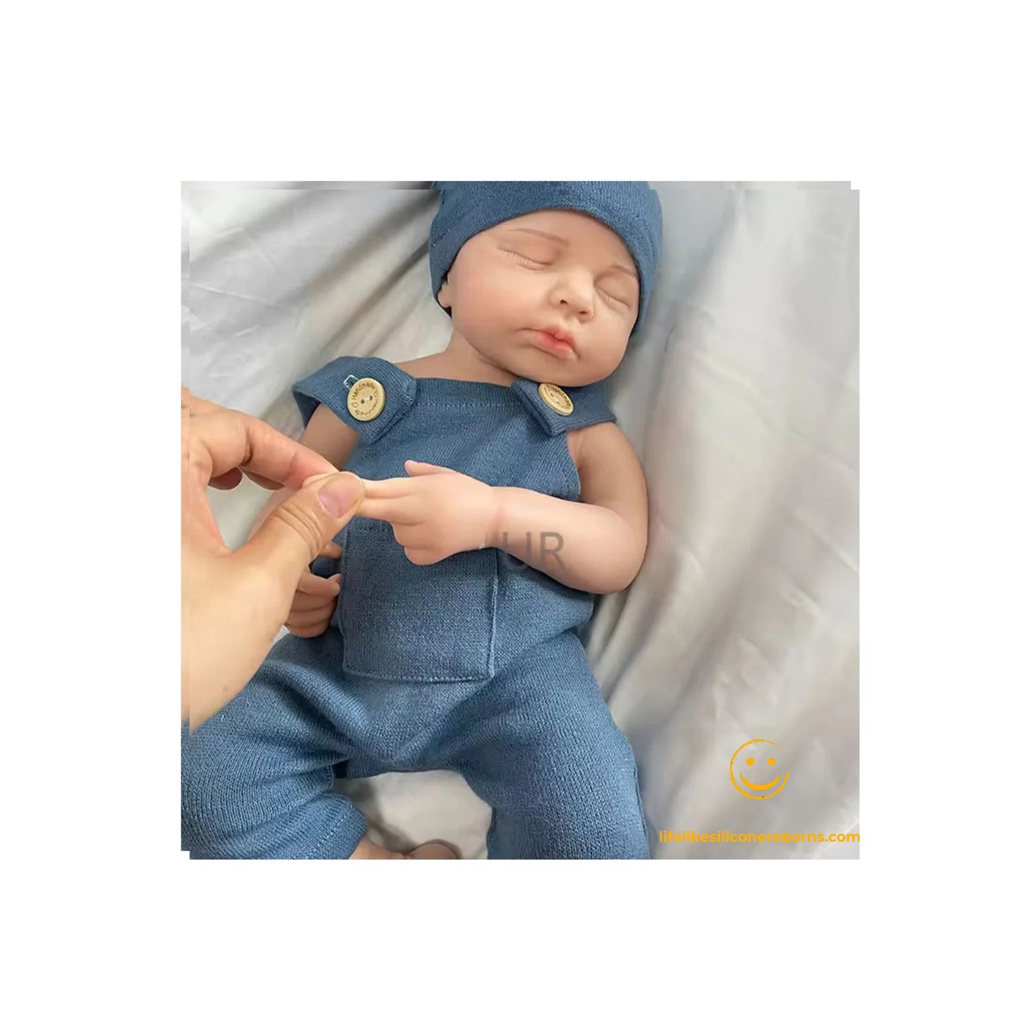 cute sleeping realistic silicone reborn baby doll in a blue outfit with a hat, lying on a white blanket.